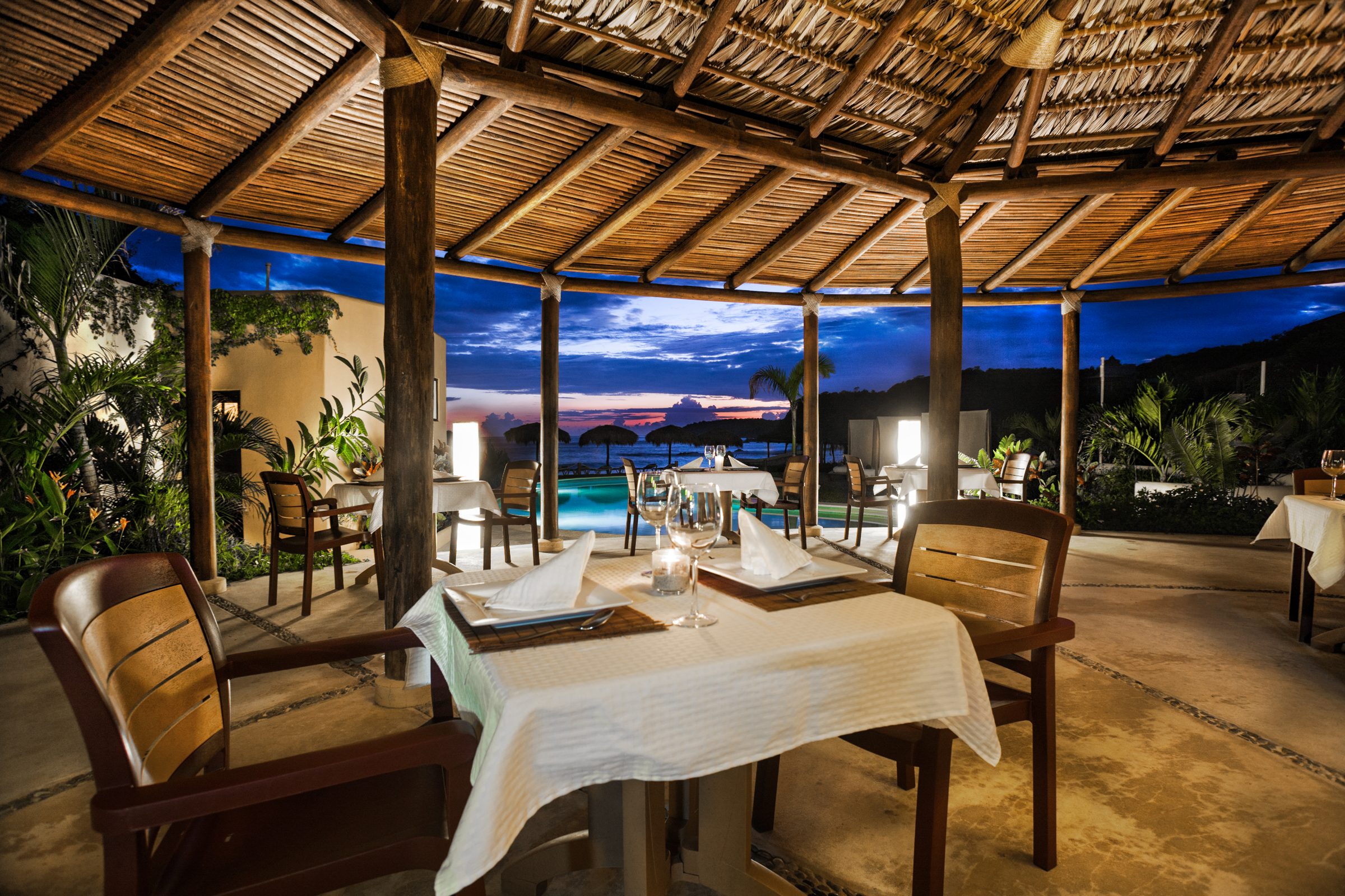 La salle à manger sous la palapa au crépuscule, tables nappées de blanc, le Pacifique au loin